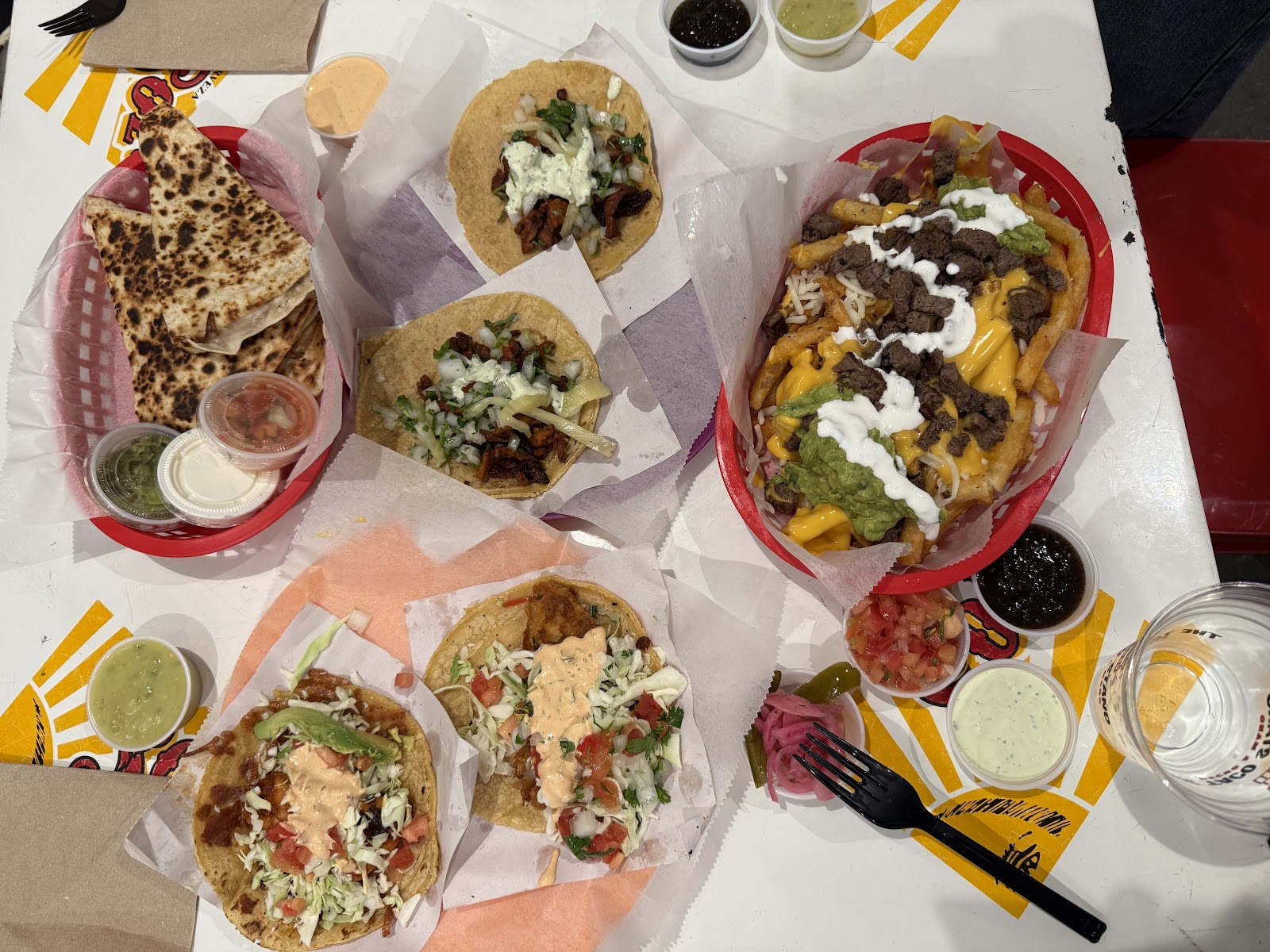An overhead spread of street tacos, a quesadilla, and loaded fries in red baskets with cups of salsa at The Taco Stand