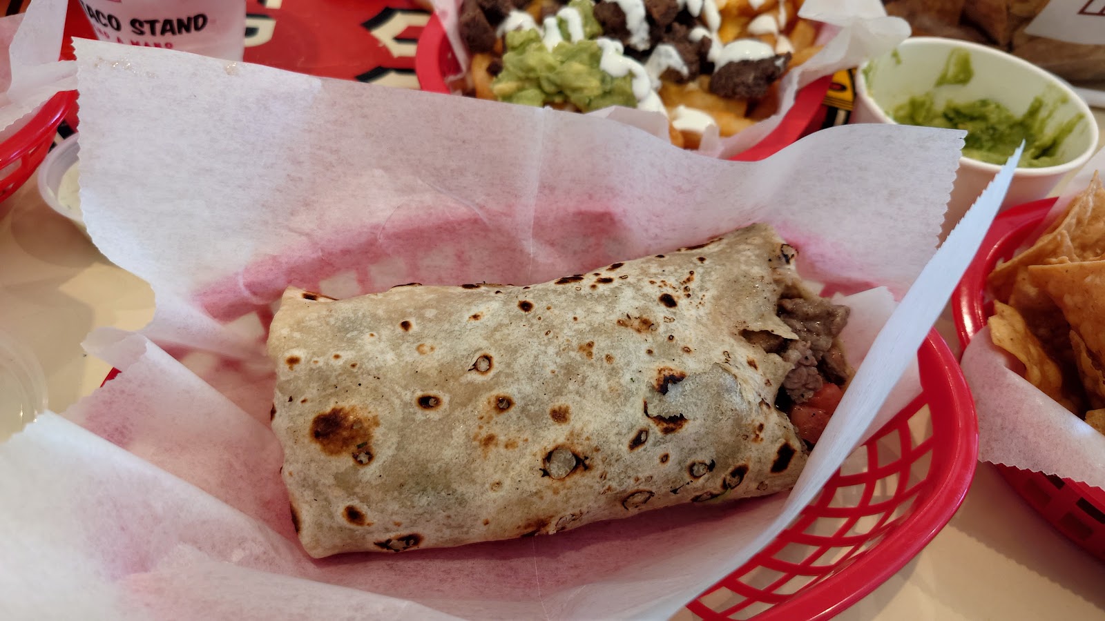 A grill-marked carne asada burrito in a red basket beside mar y tierra fries at The Taco Stand