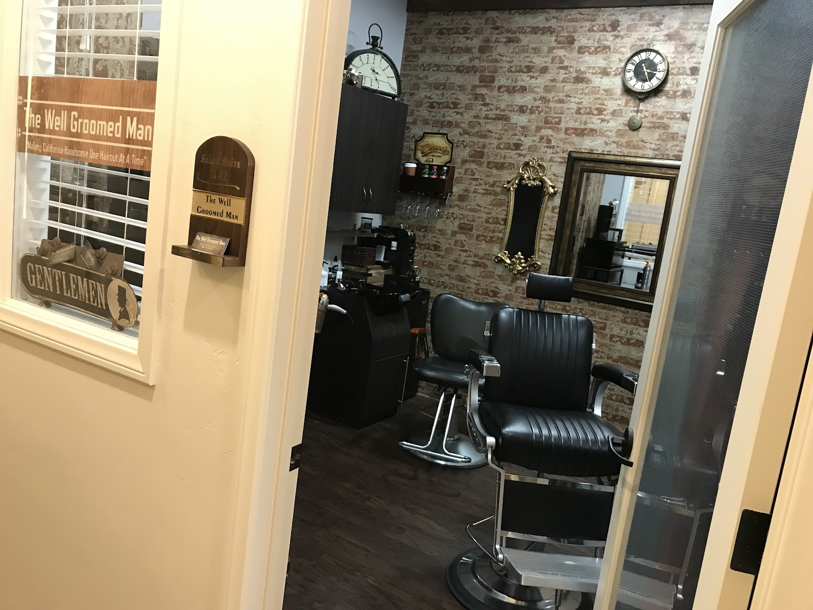 The Well Groomed Man private suite: a single black leather barber chair against exposed brick, with an ornate gold mirror and vintage clock