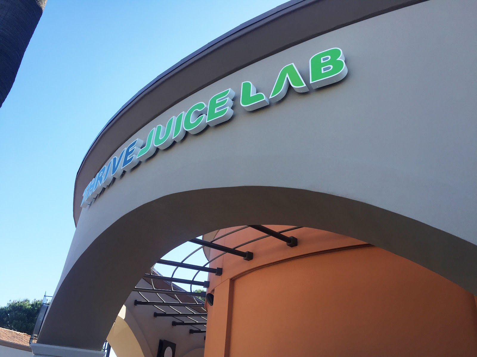 The Thrive Juice Lab storefront sign in green and teal lettering on a curved cream facade under a blue sky