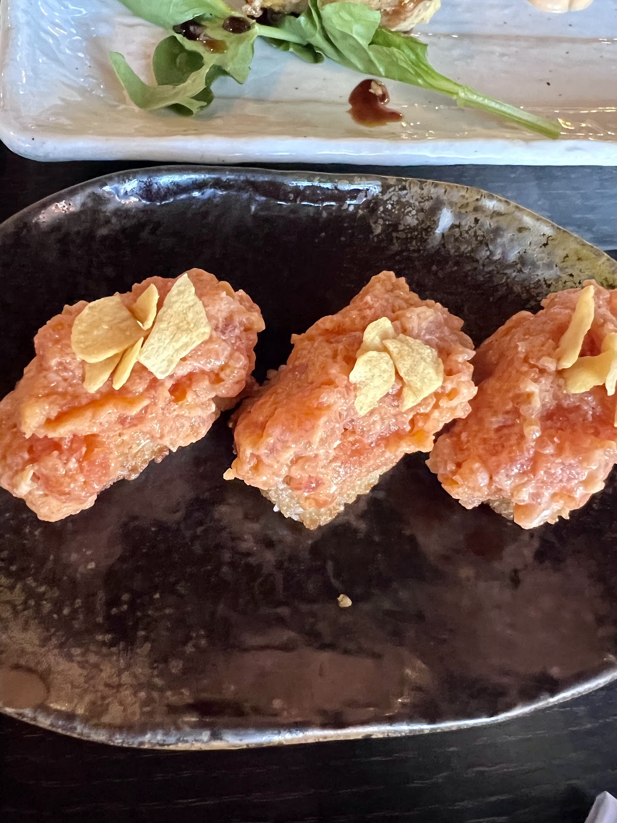 Three spicy tuna on crispy rice bites topped with garlic chips on a dark ceramic plate at Totoya