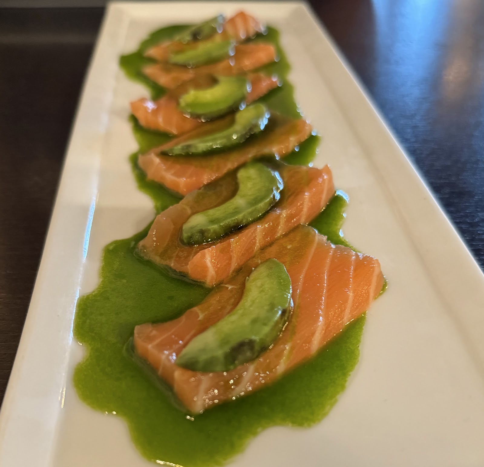 Salmon carpaccio with avocado laid out in a row over green sauce on a long white plate at Totoya