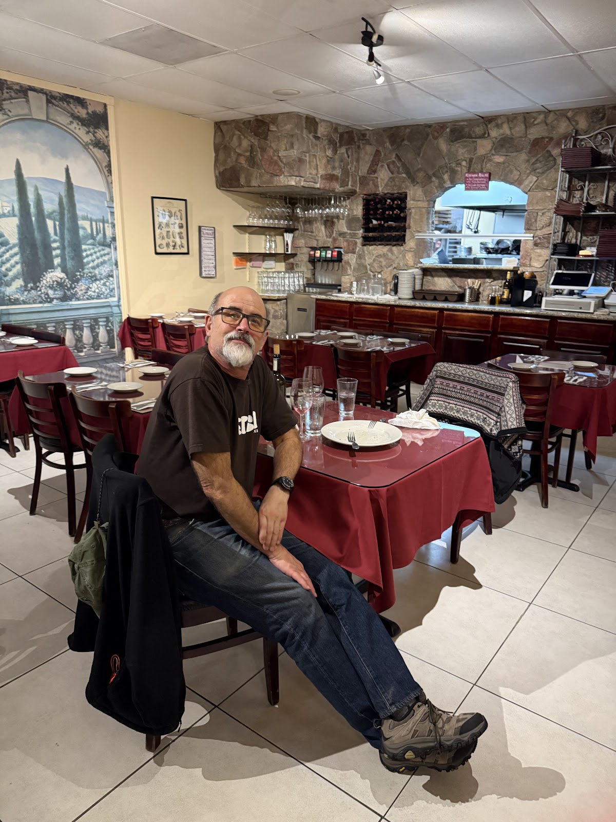 The Tutto Famiglia dining room: a painted Tuscan vineyard mural, a stone arch over the open kitchen, wine glasses on a counter, and maroon-clothed tables