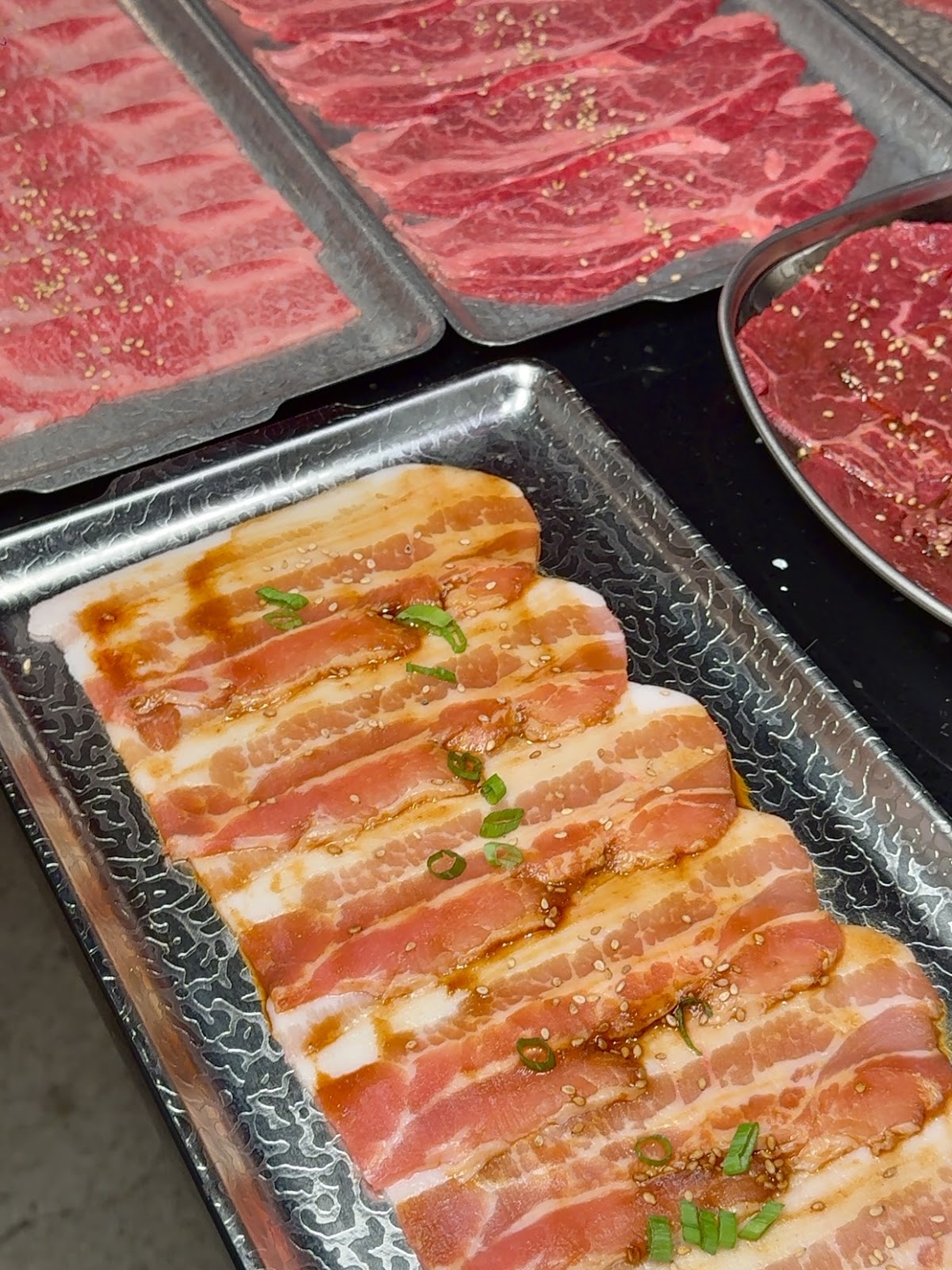 Metal trays of marbled raw beef and sauced, sesame-topped pork belly slices ready for grilling