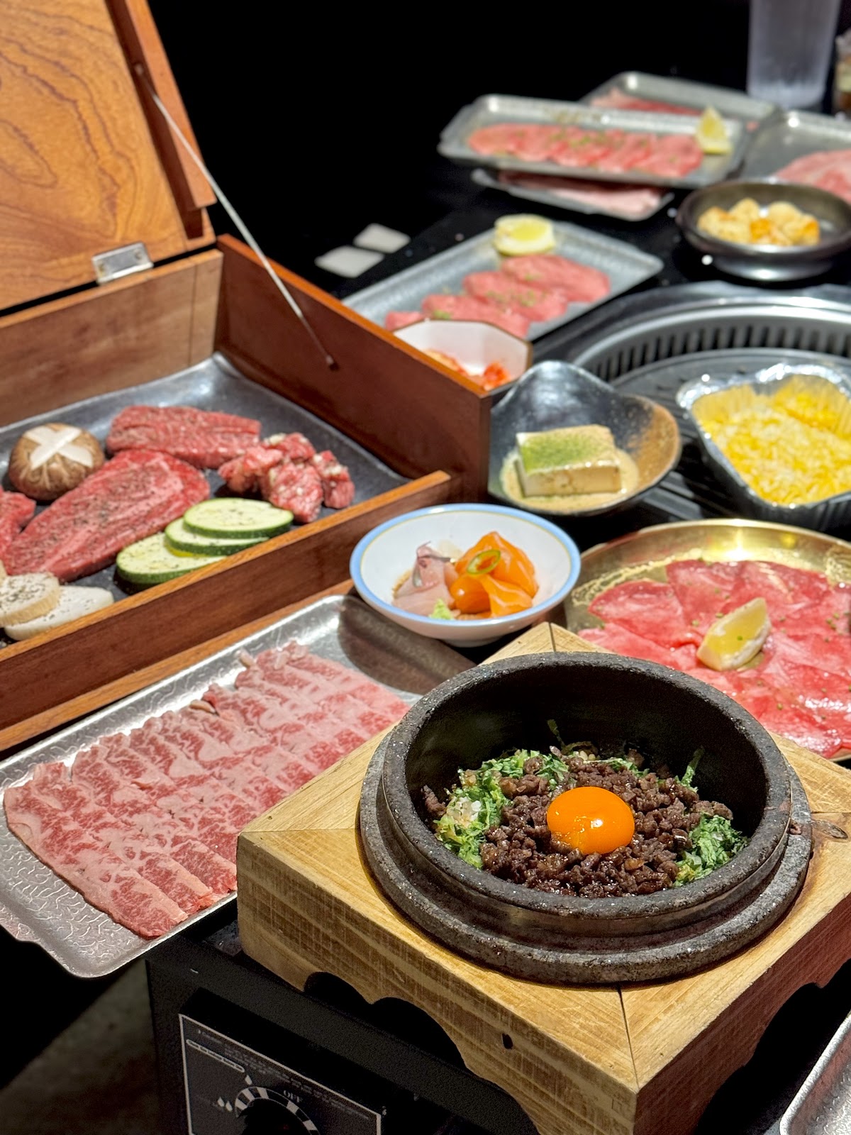 An ornate wooden presentation box of premium wagyu cuts with grilling vegetables, a stone-pot beef bowl, and sashimi on the table