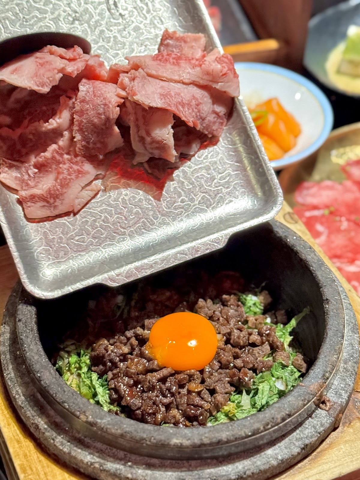 A hot stone pot of minced wagyu beef over greens with a raw egg yolk on top, beside a tray of sliced wagyu