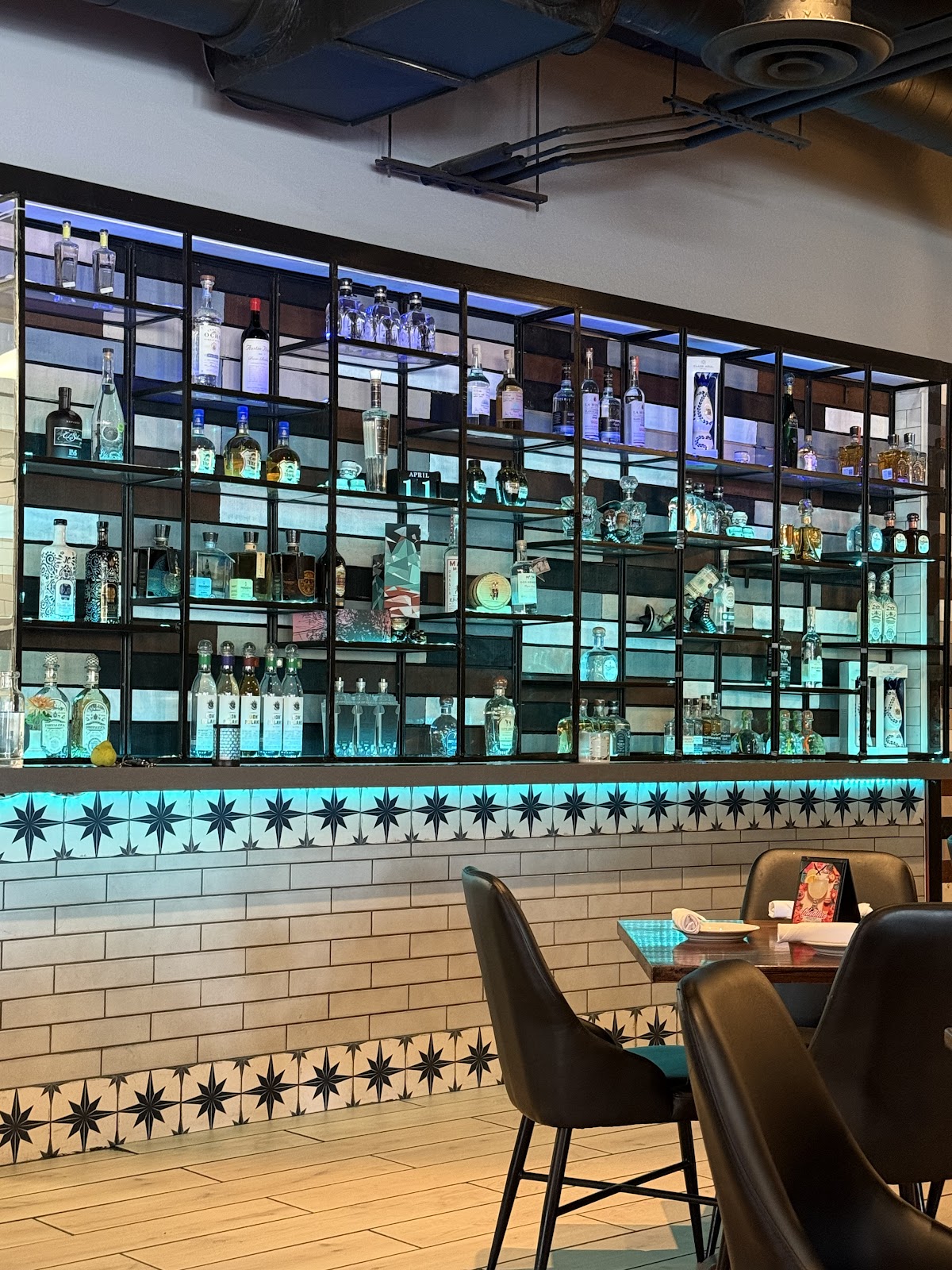 The back-lit tequila and mezcal shelf at Xclusive Mexican Kitchen glowing teal over a white subway-tile wall with an agave-tile backsplash